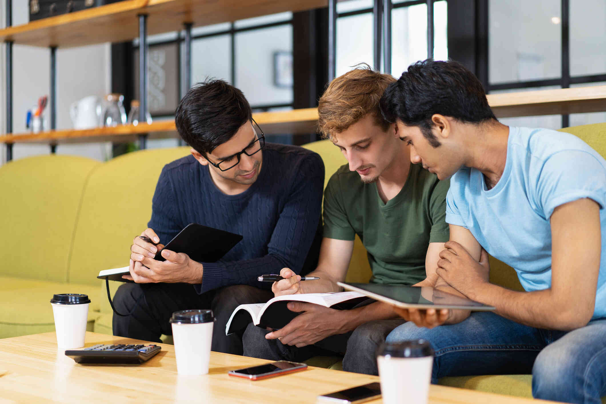 Three men sit on a couch and look down at notebooks which are spread out across their laps.